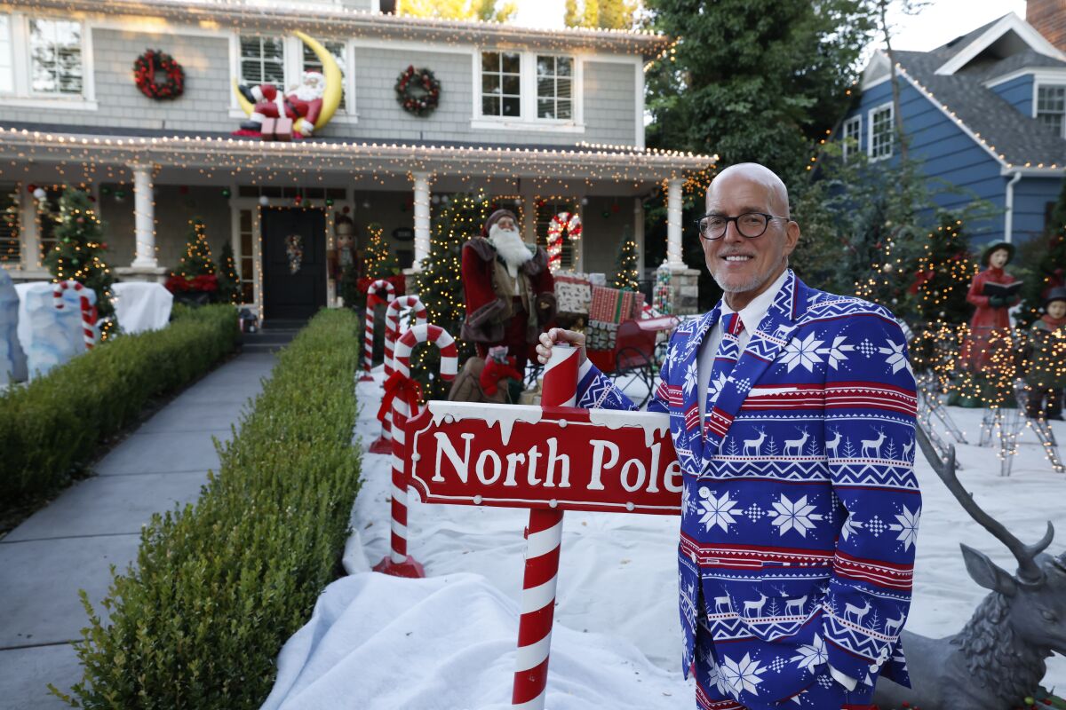 A man in a Christmas-print suit standing in front of an excessively decorated house.