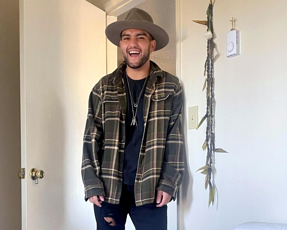 A young man in a checkered shirt and a brimmed hat smiles widely while standing in a doorframe, looking at the camera.