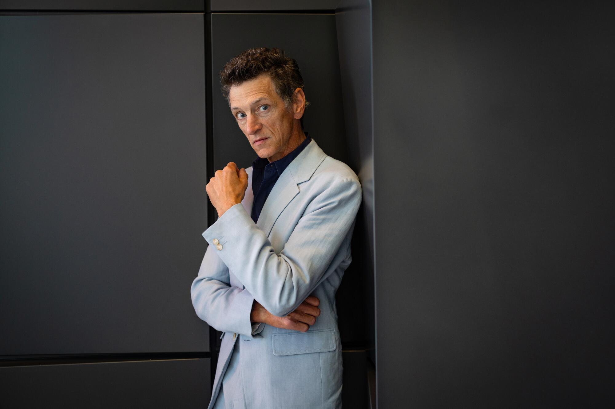 John Hawkes wears a light-colored jacket against a dark wall for a portrait.