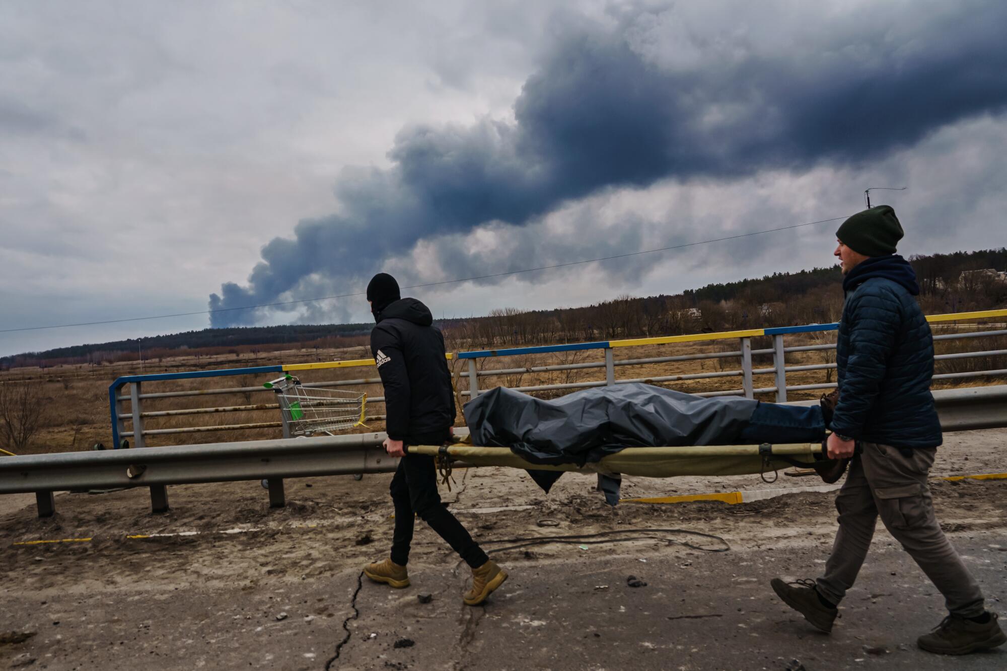 Two people carry a stretcher with a body as smoke rises in the distance.