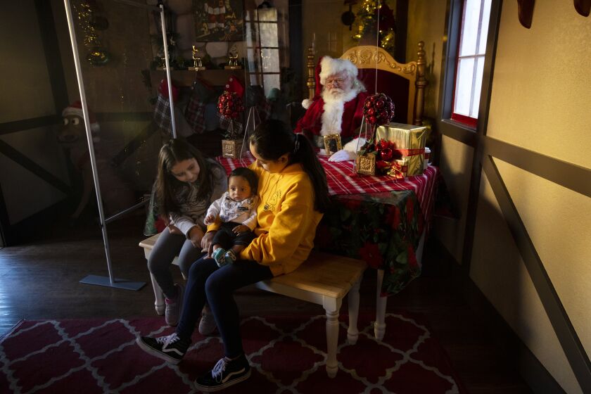 COMMERCE, CA - NOVEMBER 28: Working behind plexiglass Santa (Ray Hamlett), right, at Catherine Mejia, 7, left, Damian Rocha, 4 months, middle, and Lizzette Mejia, 11, have their photo taken inside his workshop at The Citadel Outlets on Saturday, Nov. 28, 2020 in Commerce, CA. During the coronavirus pandemic shoppers gather at The Citadel Outlets in Commerce, CA. There are new Los Angeles County COVID-19 restrictions set to take effect on Monday that would further limit public and private gatherings. (Francine Orr / Los Angeles Times)