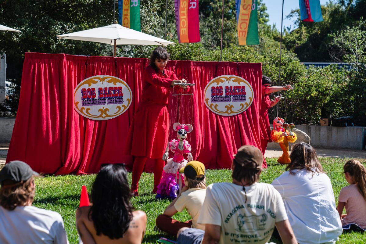 Um marionetista vestido de vermelho atua com um fantoche de rato na frente de uma multidão em um parque.