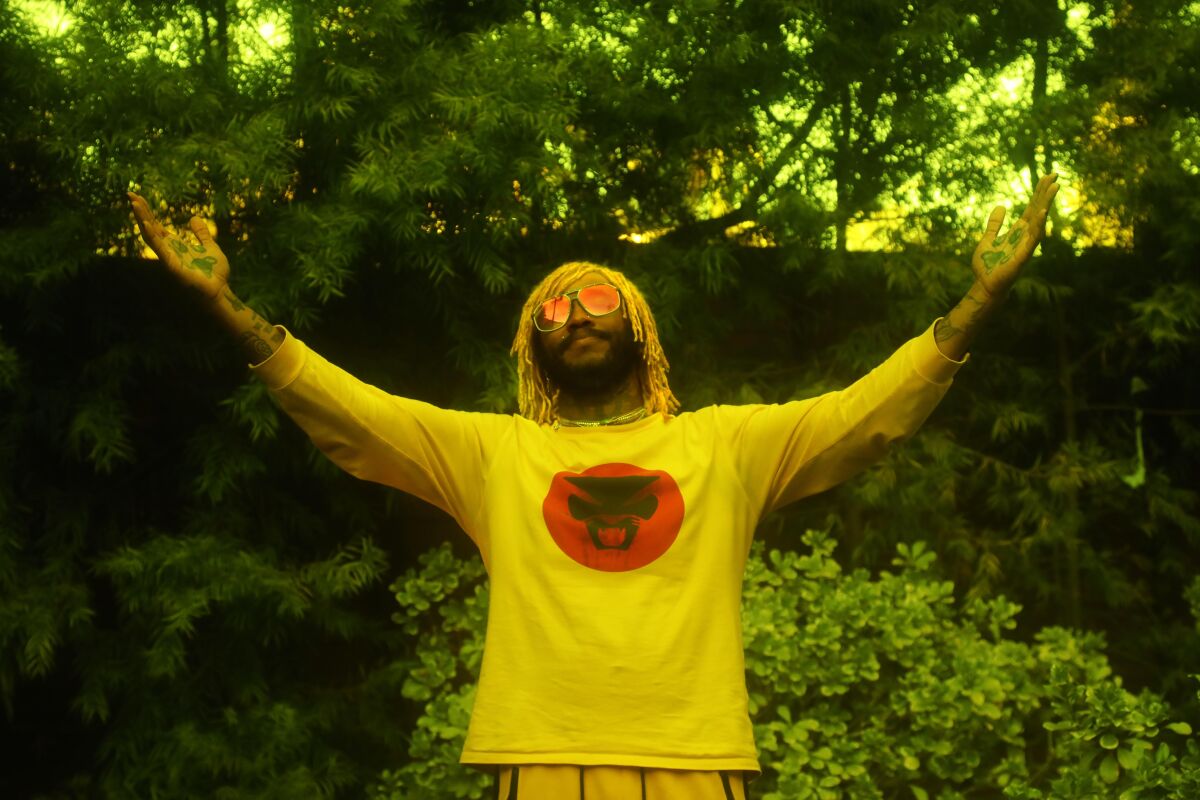 Musician stands before a backdrop of trees and shrubs, wearing sunglasses and lifting both arms.