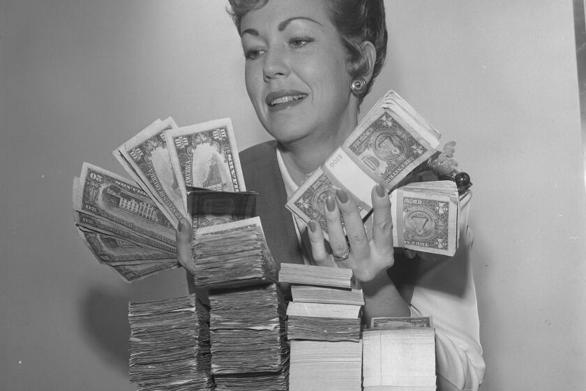 A woman holds dollar bills, 10s and 20s in front of four small stacks of cash.