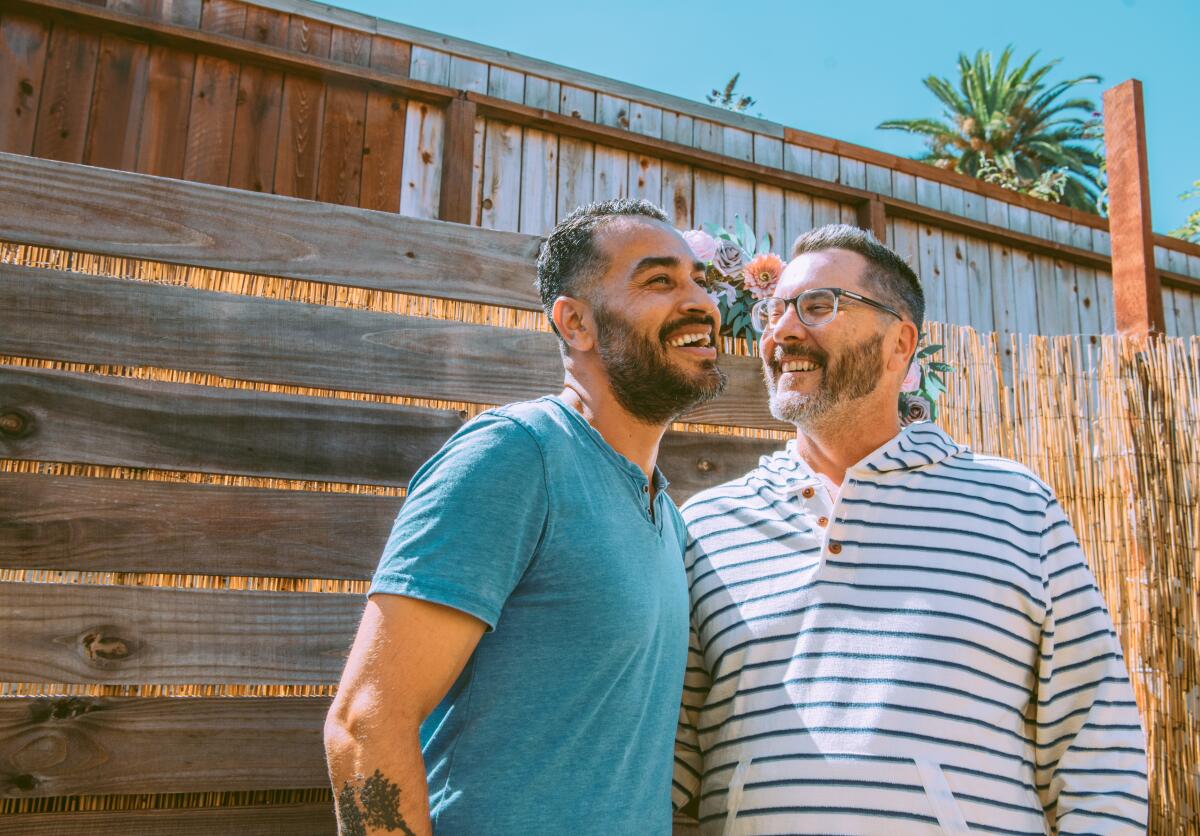 Two men stand outside in front of a wooden fence.