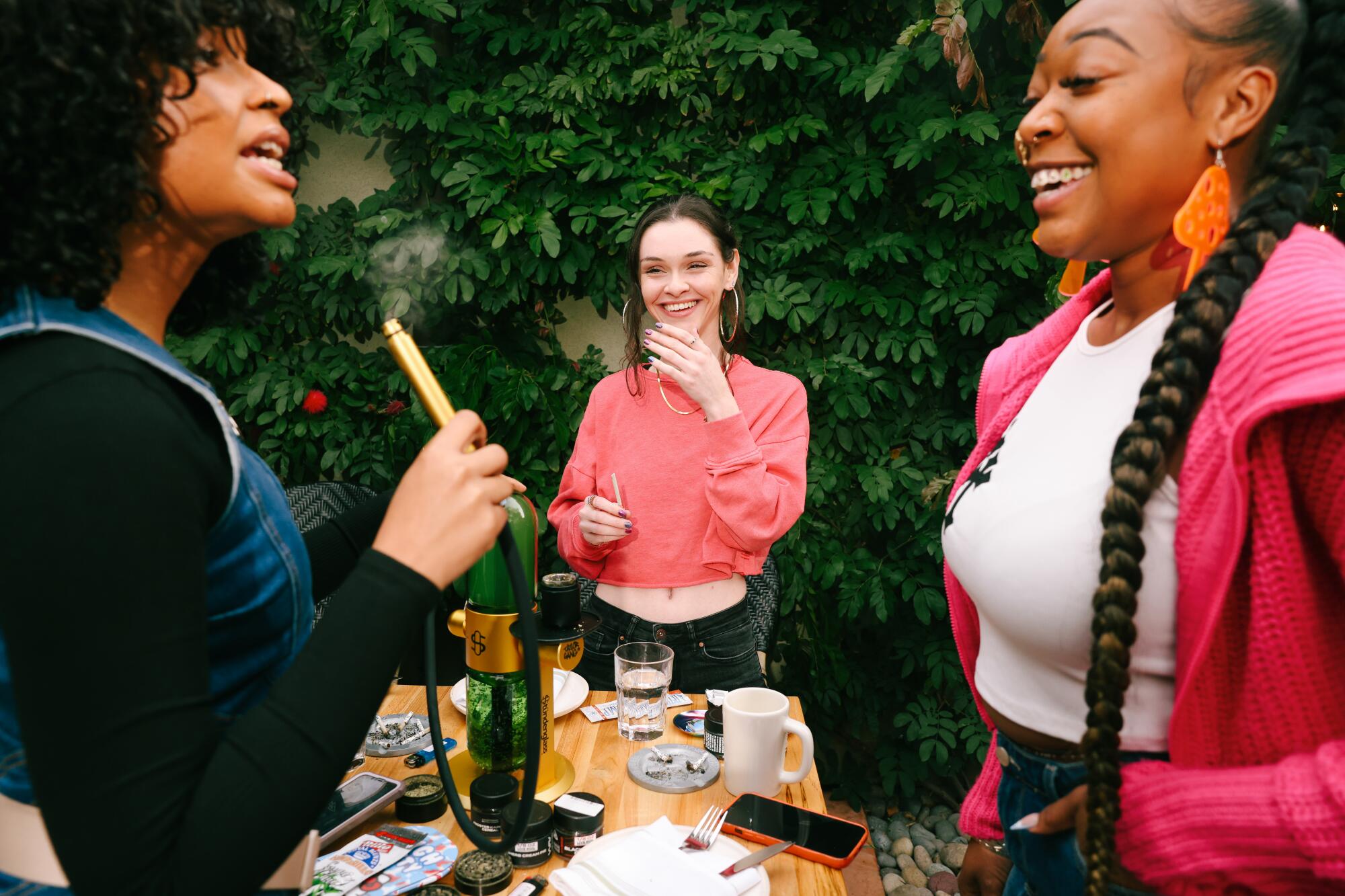 Three women laughing, one holding a bong mouthpiece and another holding a joint, at a table covered with cannabis products.