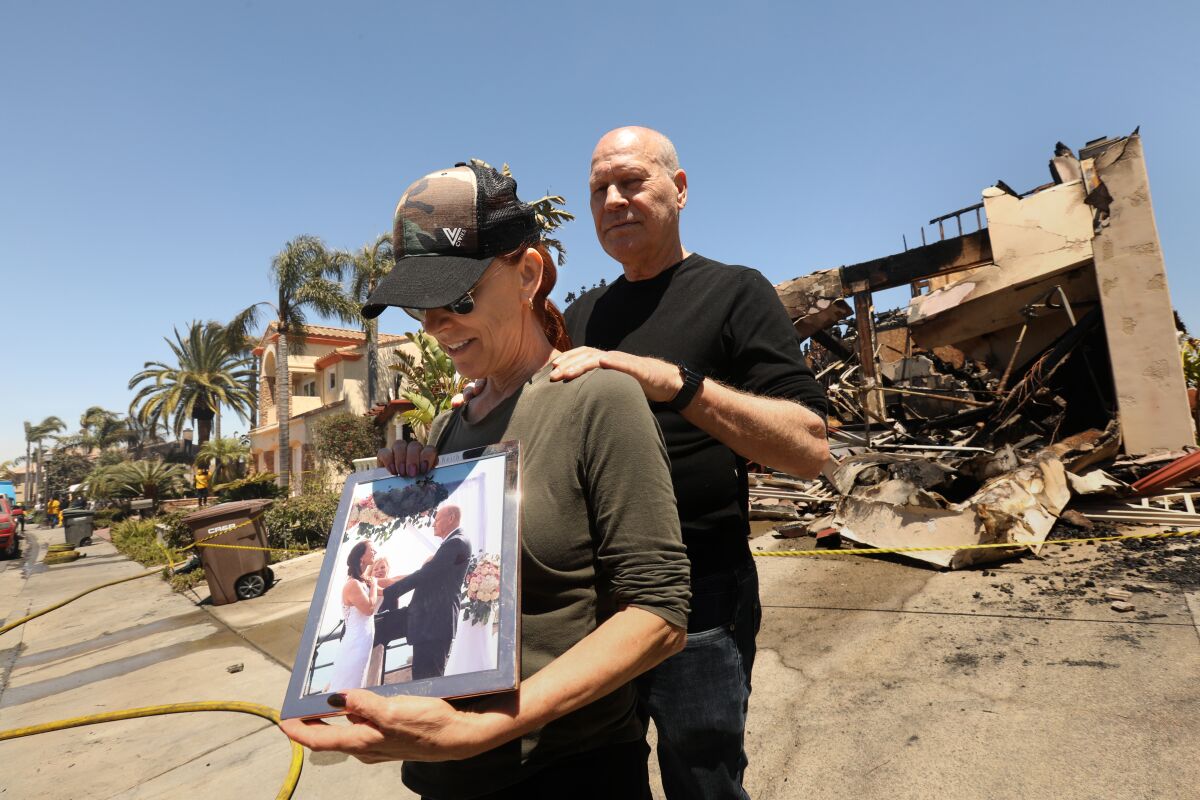 A couple stands with a framed photo in front of a destroyed house