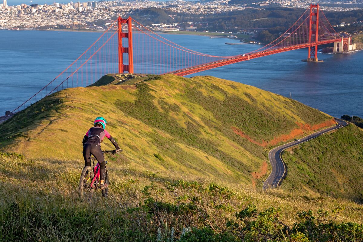 A bicyclist in a helmet rides on a grassy hill. In the distance is a stunning orange bridge, a bay and a city beyond.