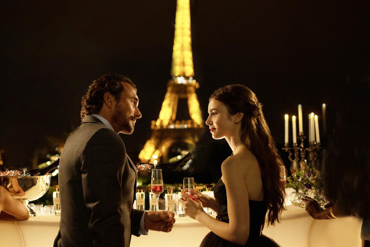 A man and woman carry drinking glasses with the Eiffel Tower in the background at night in "Emily in Paris"