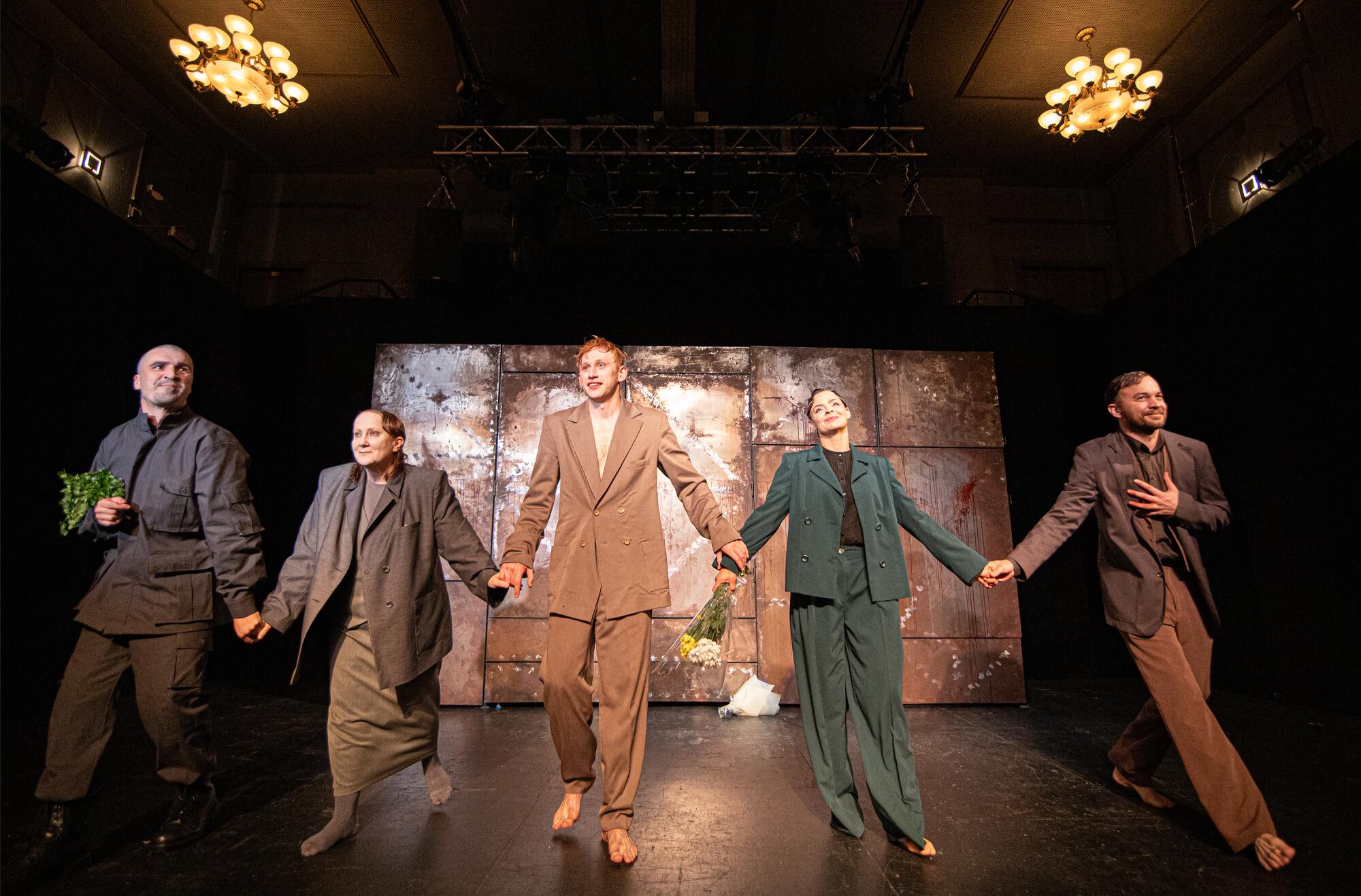 Five actors in loose-fitting suits hold hands on stage 