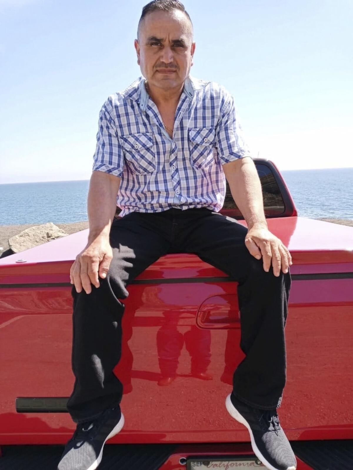 A man in a short-sleeve black-and-white plaid shirt and dark pants sits, with hands on his knees, in the back of a truck