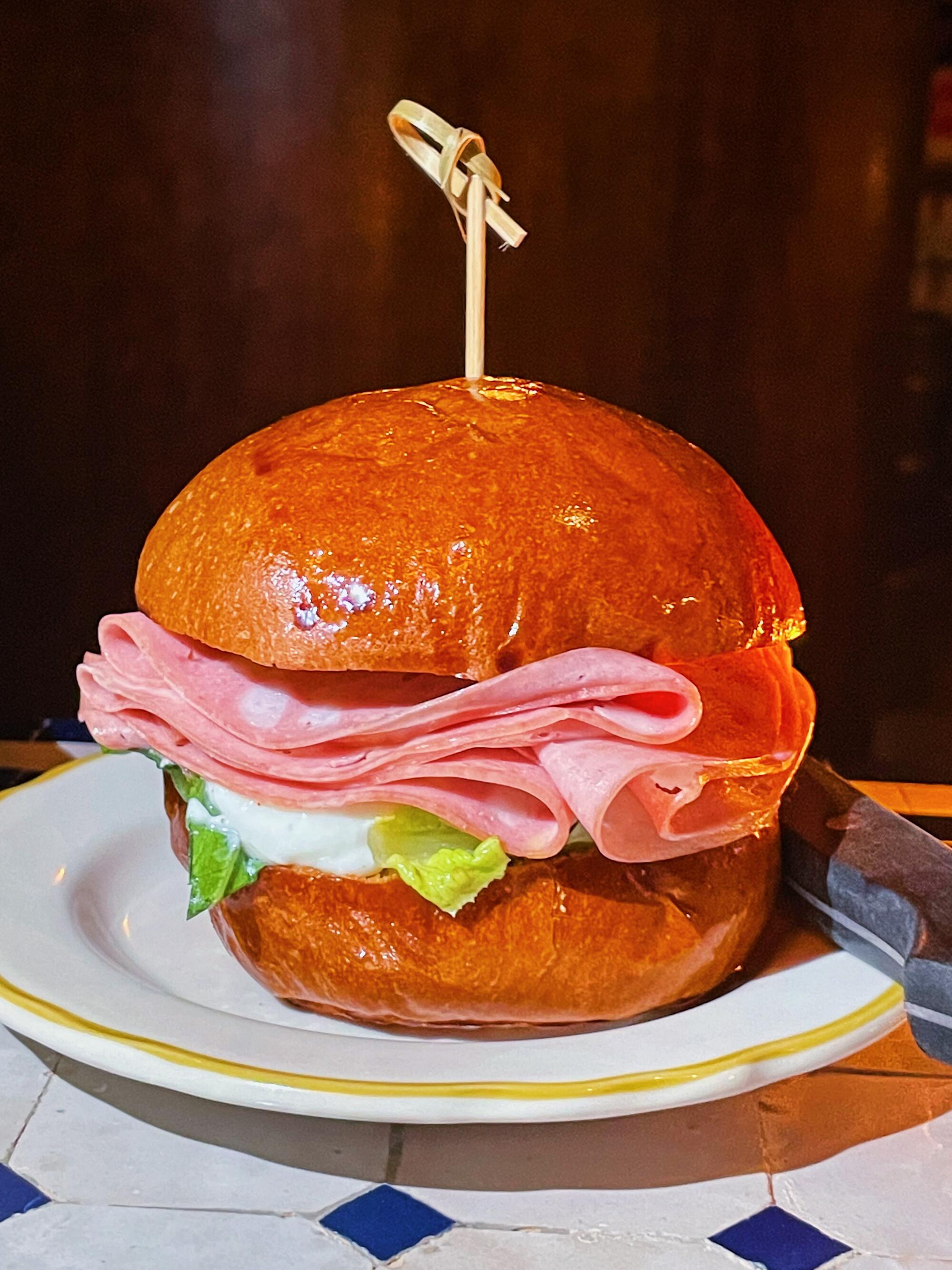 A glossy-bunned mortadella sandwich atop a blue-and-white tile table.