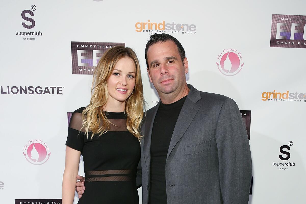 A blonde woman in a black dress stands with a man in a gray blazer