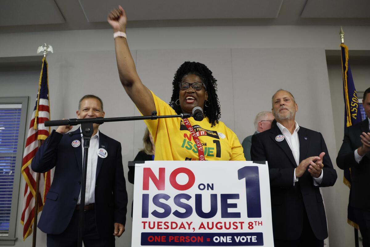 A woman speaks into a mic and raises her arm behind a sign that reads "No on Issue 1" as men applaud behind her.