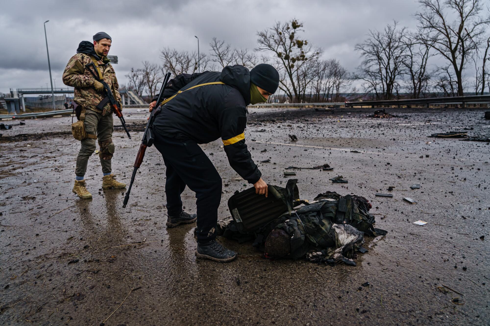 A Ukrainian soldier leans over the body of a Russian soldier while a second Ukrainian stands nearby. 