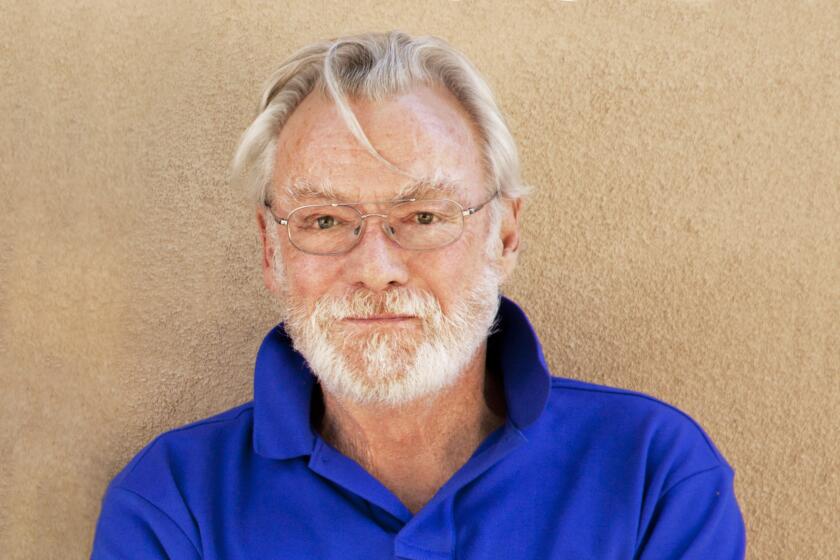 John Morris wears a blue polo with an orange insignia as he poses for photos in front of a beige wall