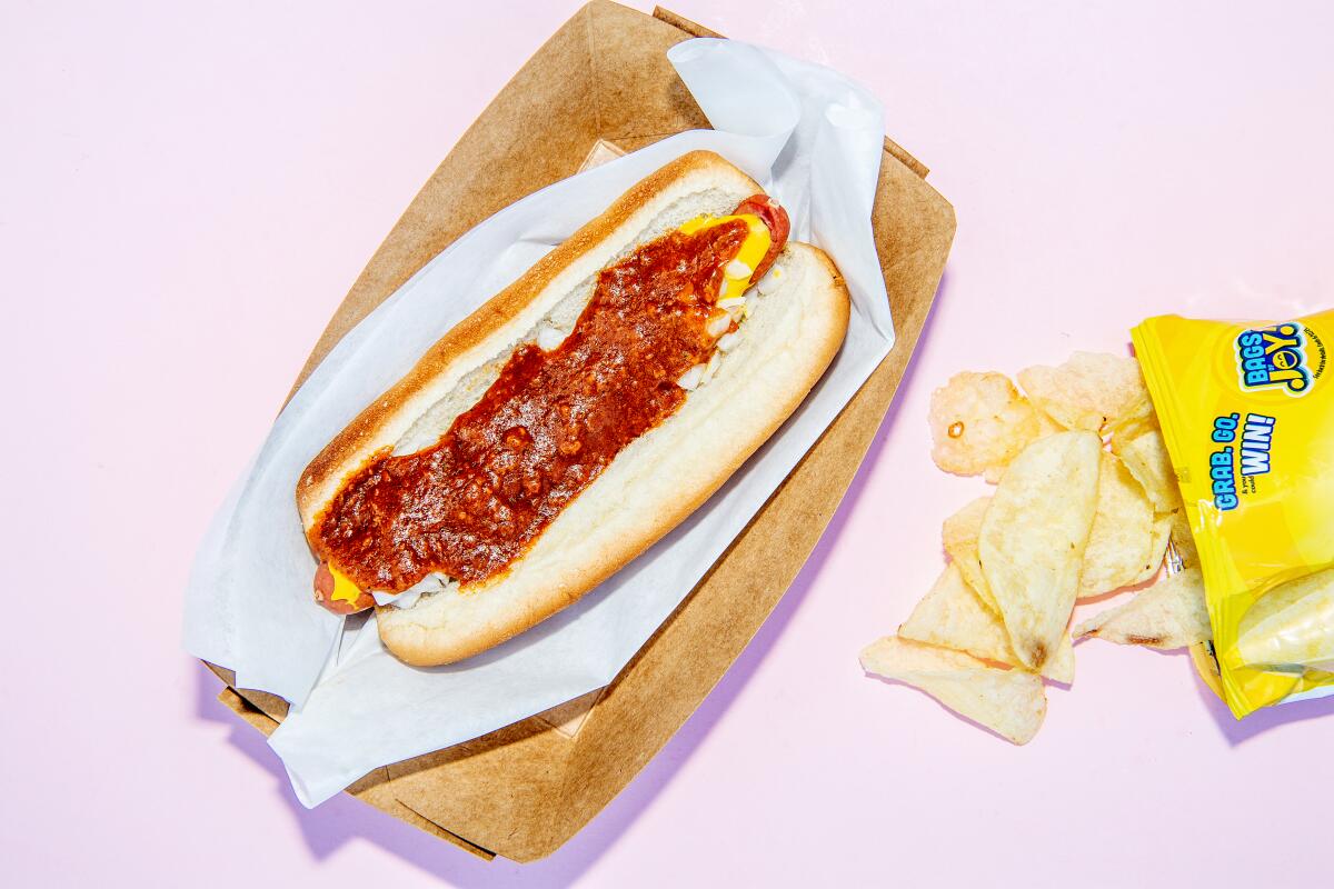 A chili hot dog on a paper wrapper next to an open bag spilling out potato chips