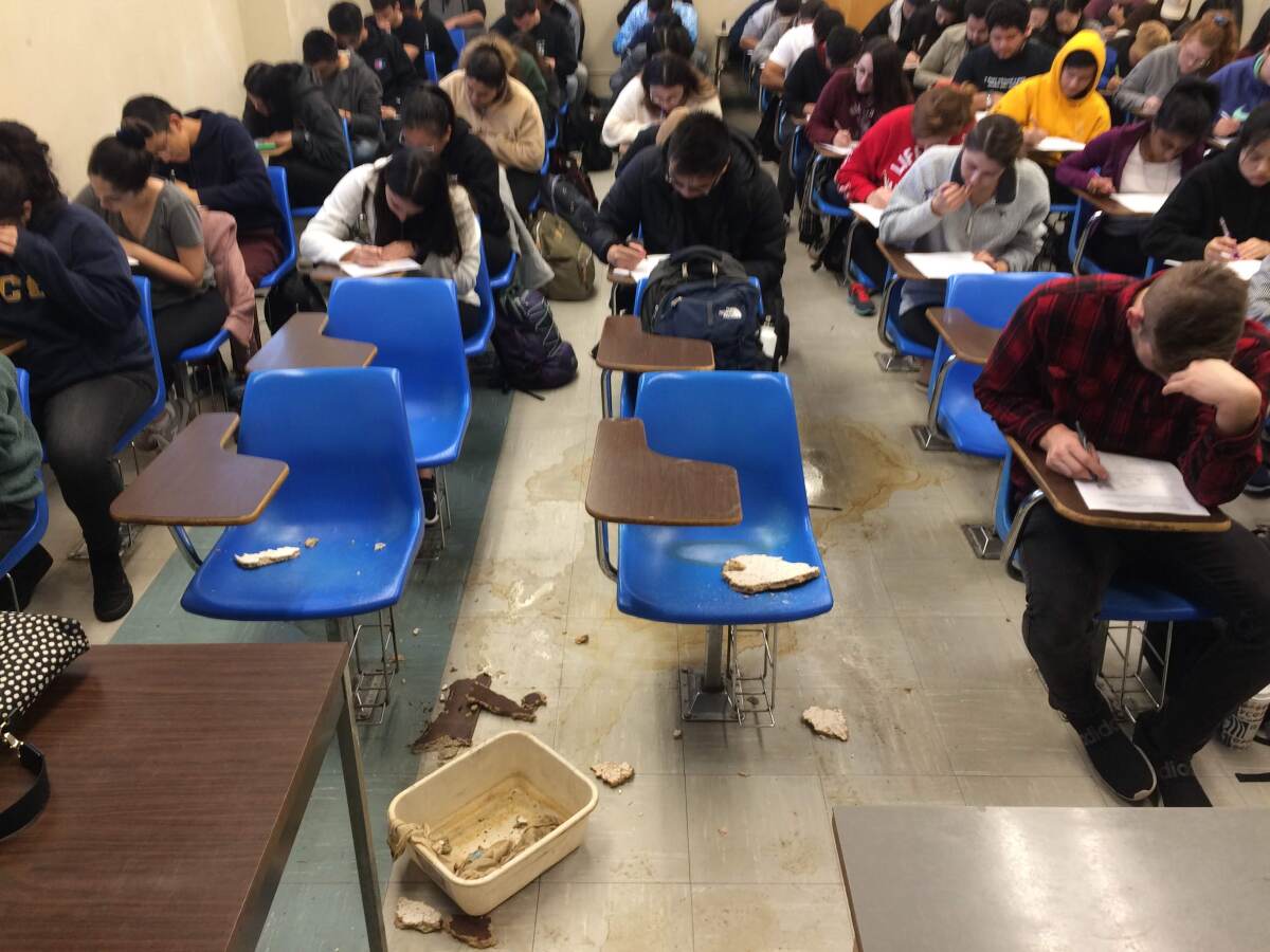 Broken pieces of ceiling tiles and water stains cover empty desks in the middle of a crowded classroom
