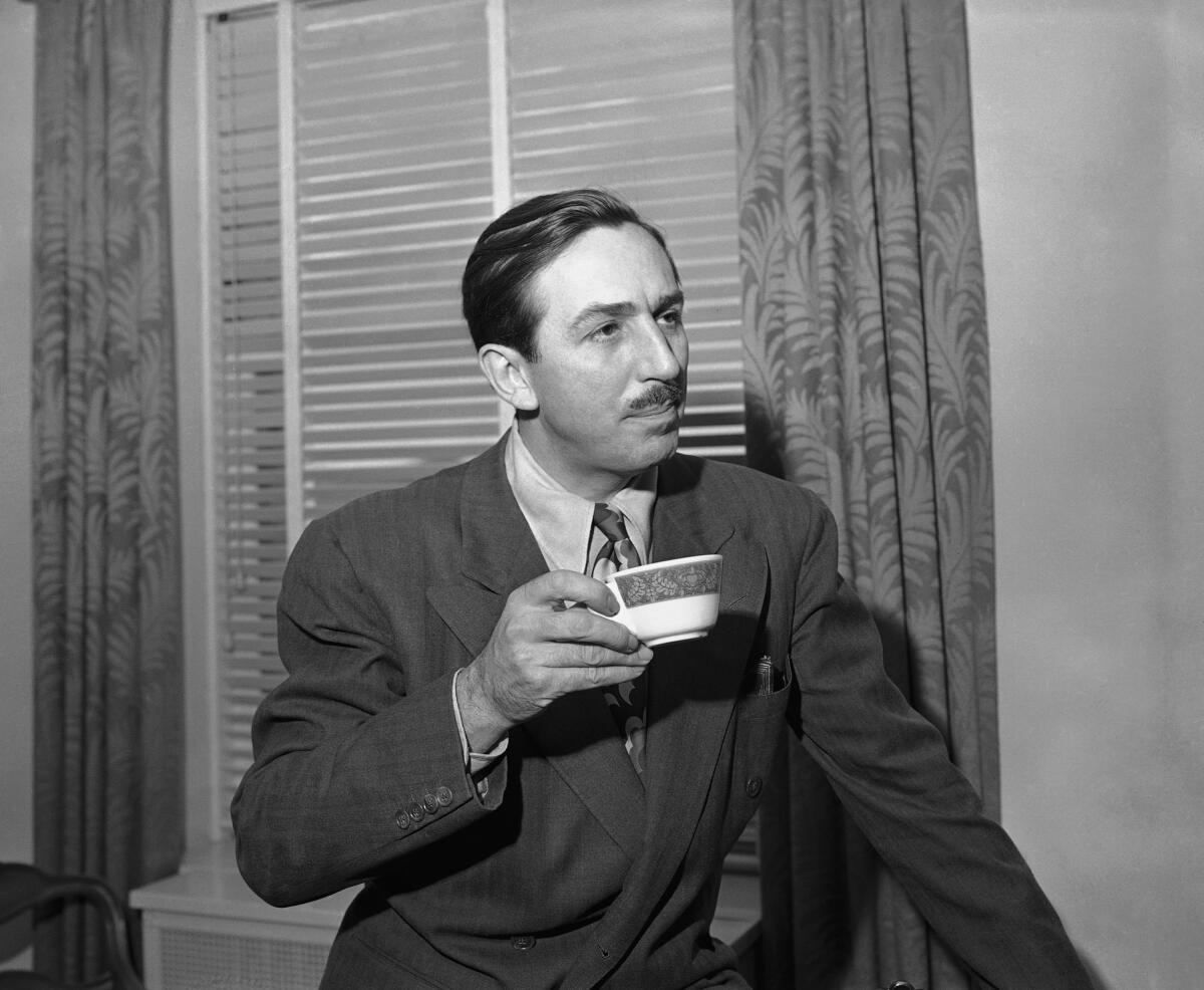 Walt Disney looks off to the right  while holding a cup in his right hand. 