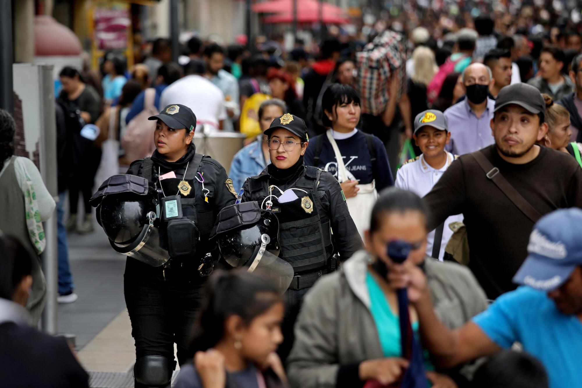 Two female Mexico City police officers in body armor walk along a crowded avenue.