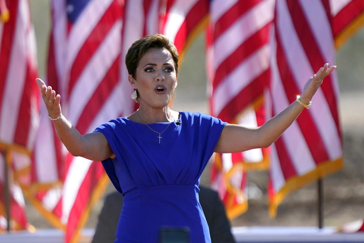 A woman in a dark blue dress, standing in front of American flags, holds her arms up and speaks. 