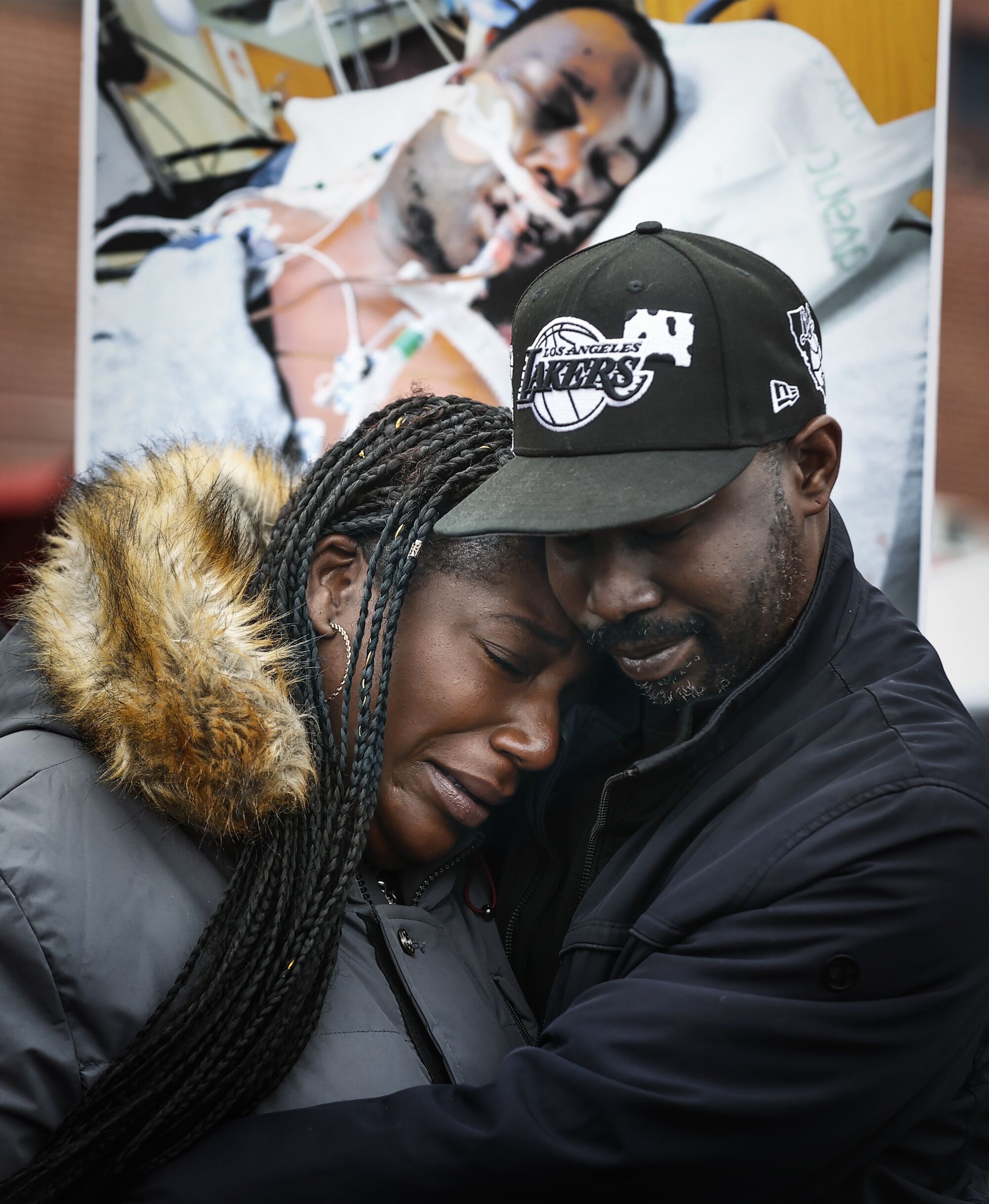 A man with his arms around a weeping woman in front of a poster of an injured man in a hospital bed