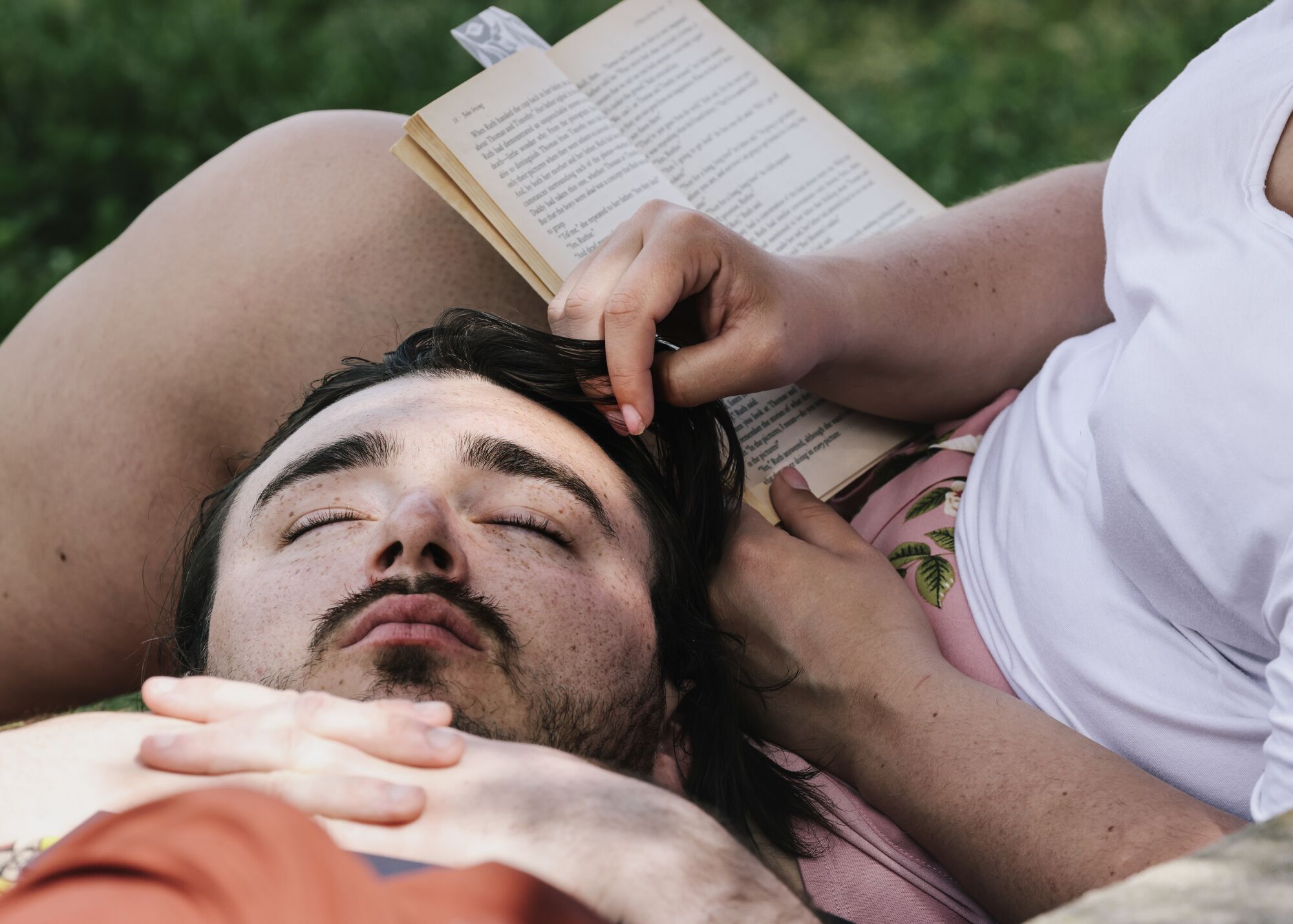 Leaning back, next to an open book, a man rests his head on the lap of another person; his eyes closed.