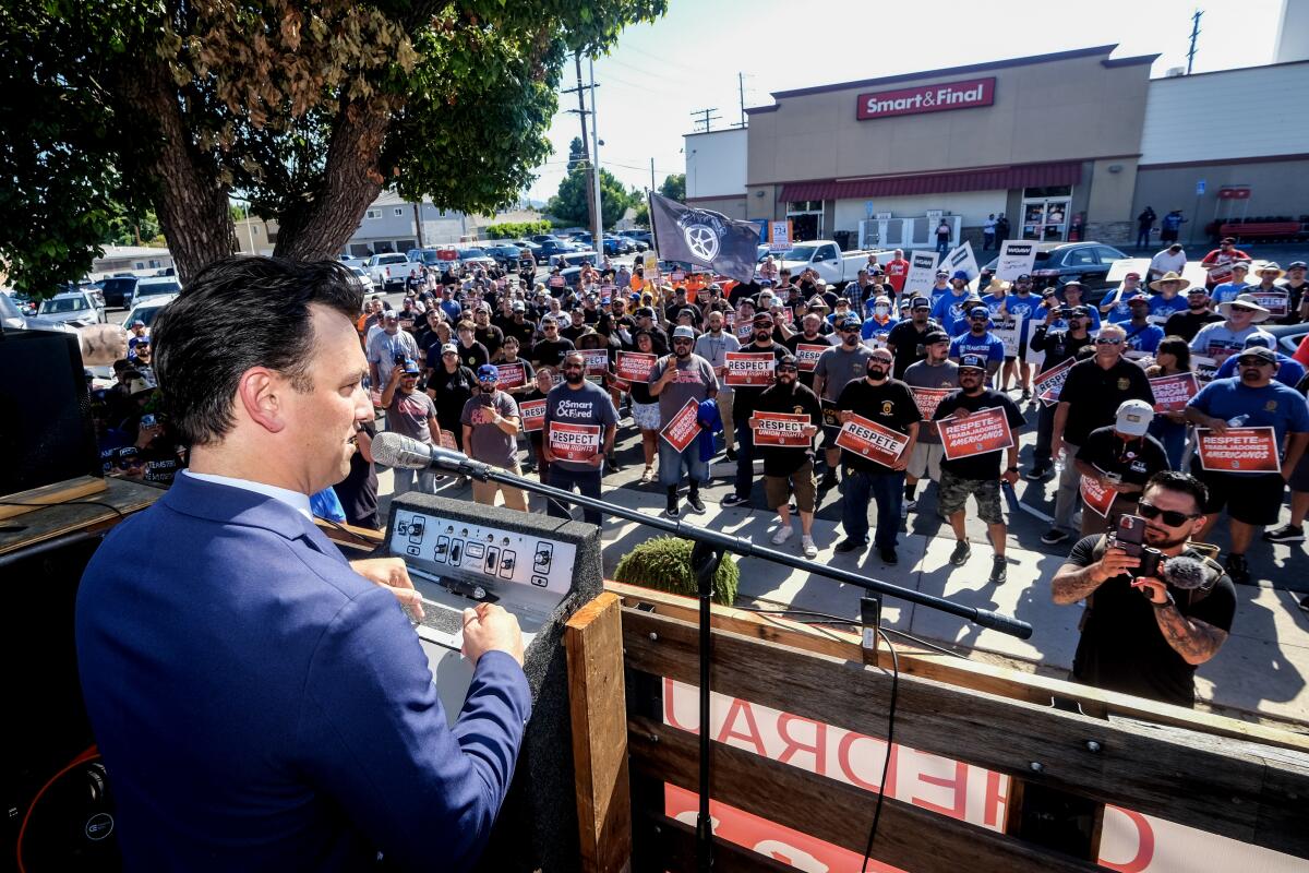 A man in a suit speaks into a microphone in front of a crowd of people with rally signs outside a store.