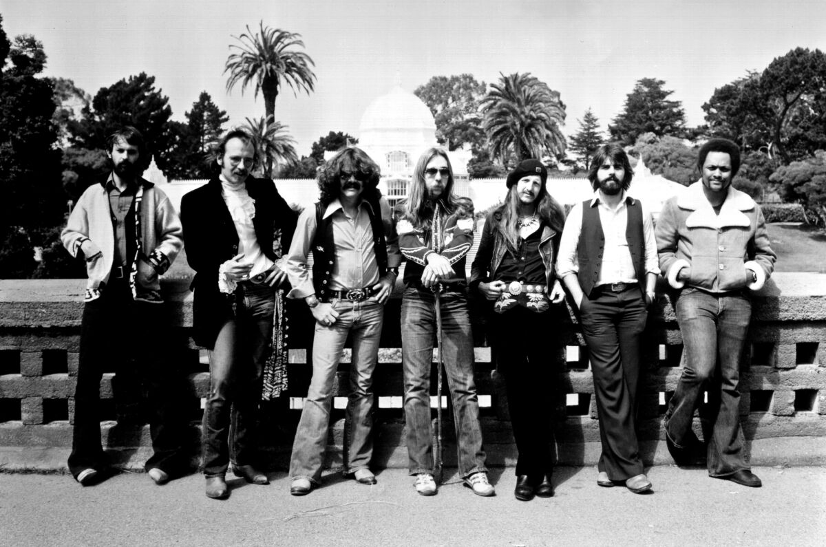 A seven-member rock band stands in front of a half wall in the 1970s