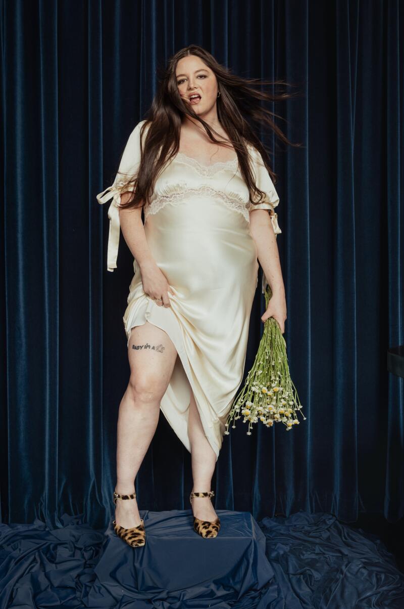 A woman, holding a green bag with her left hand, raises the hem of her dress to reveal her right leg while posing for a photo