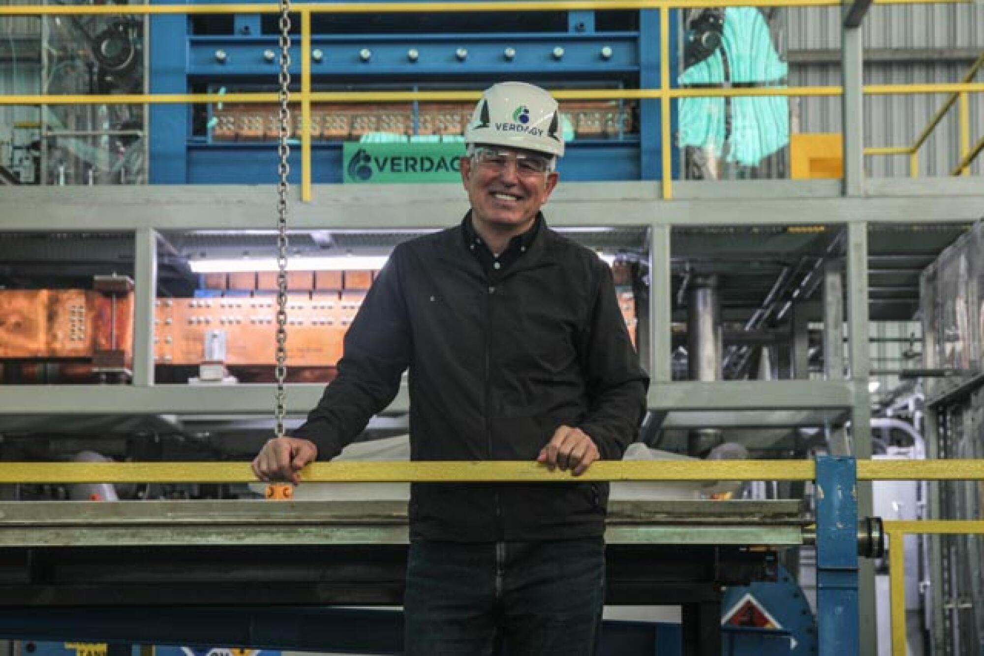 A man wearing a black shirt and a hard hat stands in an industrial setting.