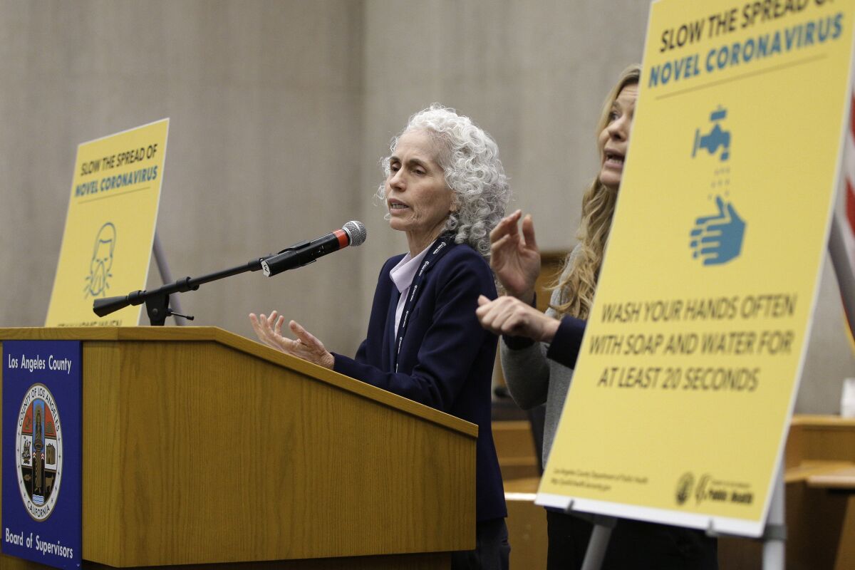 Barbara Ferrer speaks at a lectern next to yellow signs advising the use of masks and frequent hand-washing