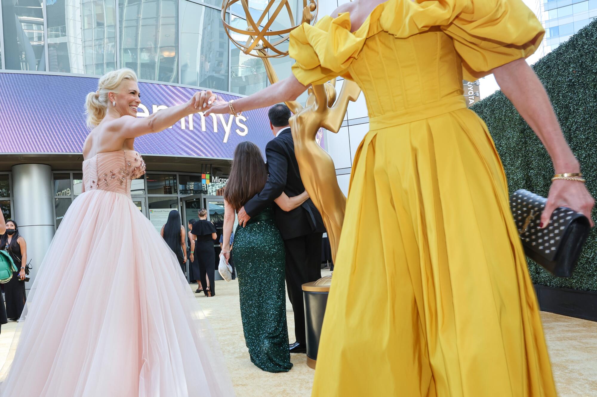 A woman in a glamorous pink dress reaches out to take the hand of a woman in a yellow dress.