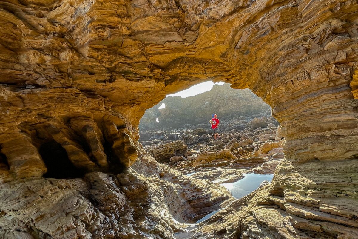 Framed by a rock arch, a person walks along shoreline rocks.