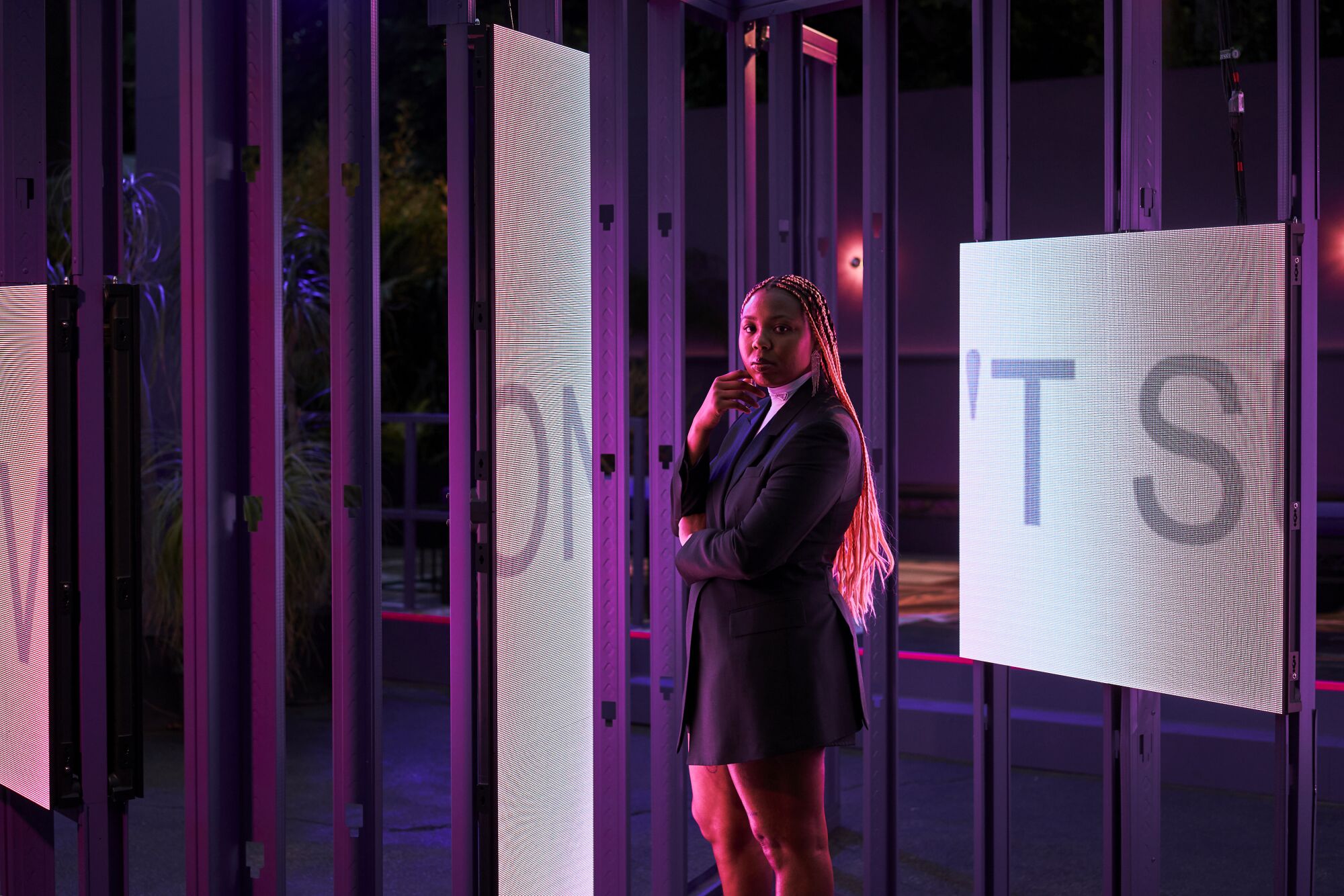 A woman with pink braids stands in front of screens projecting letters