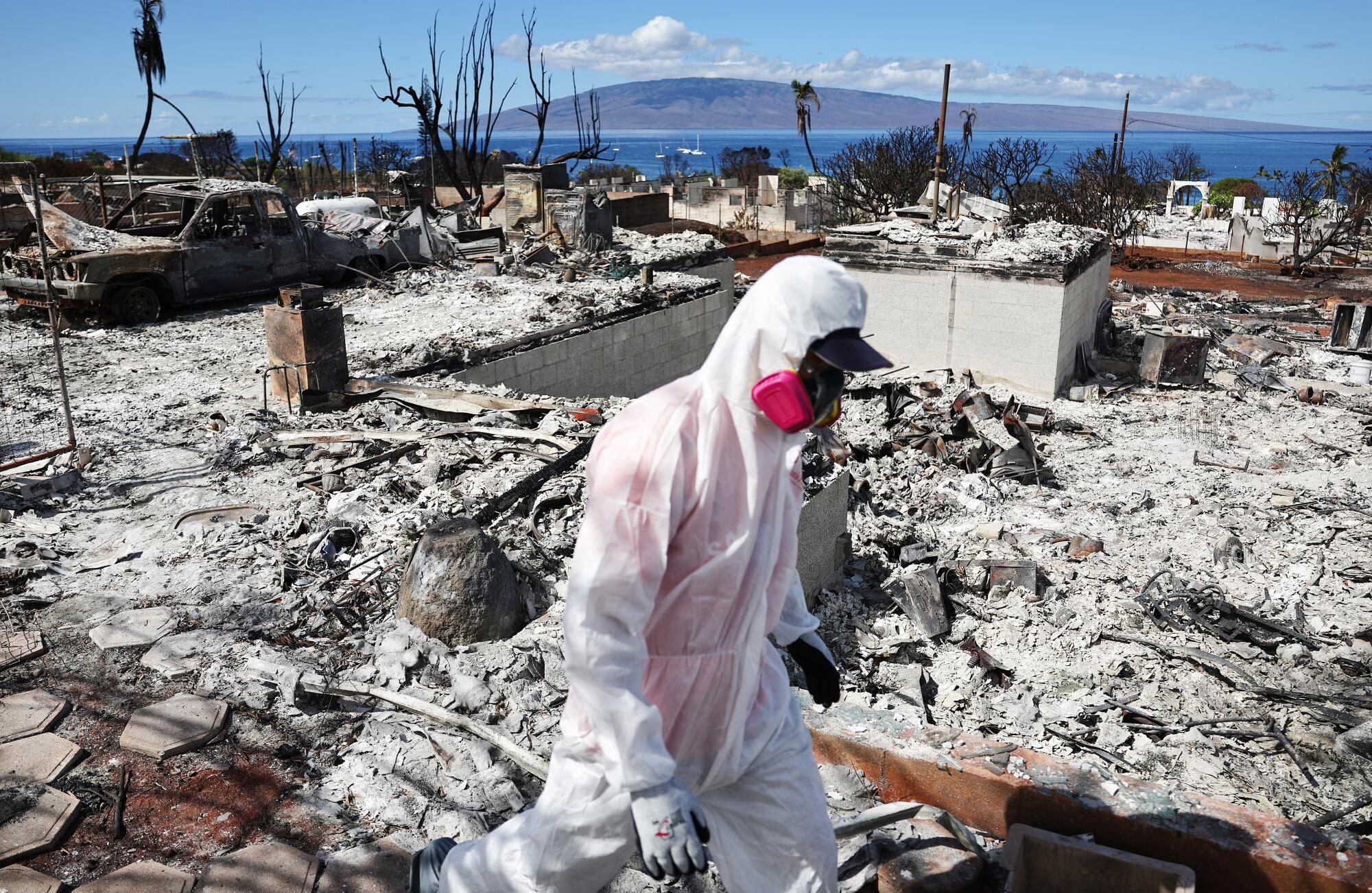 A volunteer in protective gear walks through a burn site in Lahaina, with the ocean in the background.