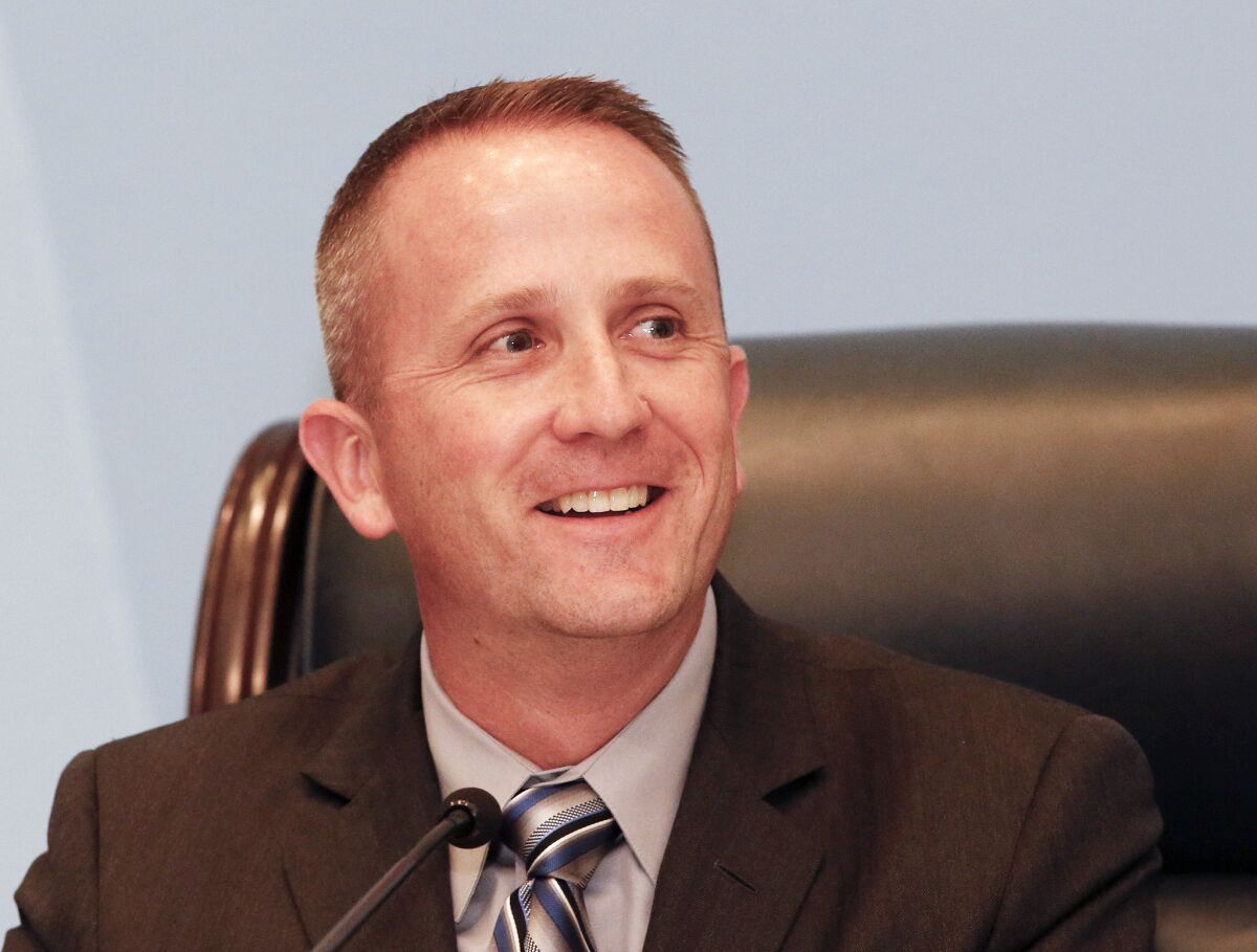 Lou Vince smiles wearing a coat and tie and sitting in front of a microphone