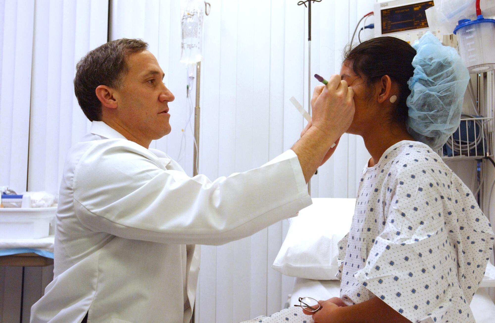 A doctor in a white lab coat marks on the face of a woman in a hospital gown and surgical cap.