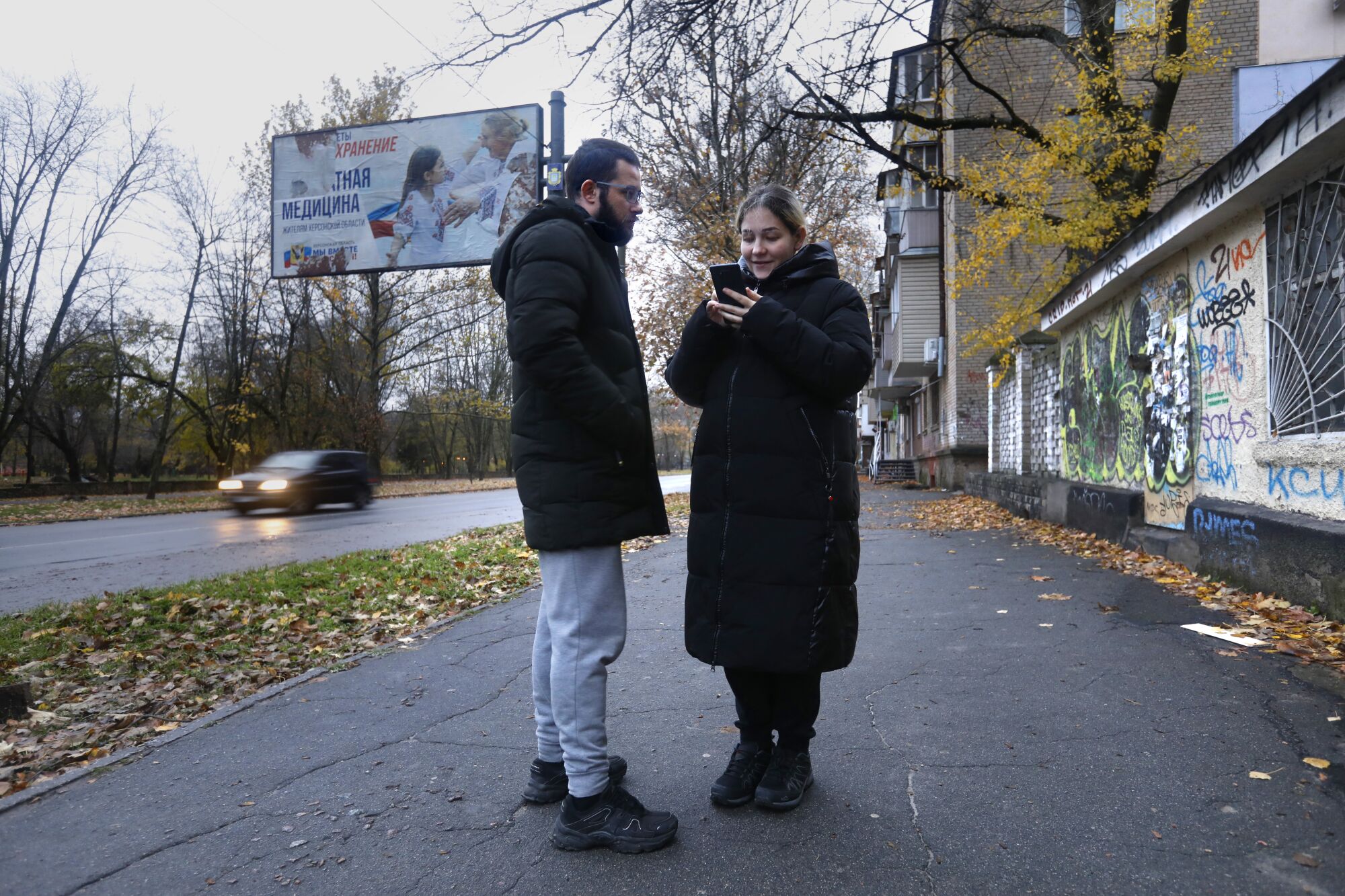 A bearded man, left, and a woman holding a cell phone, both in dark winter jackets, stand beside a road near buildings 