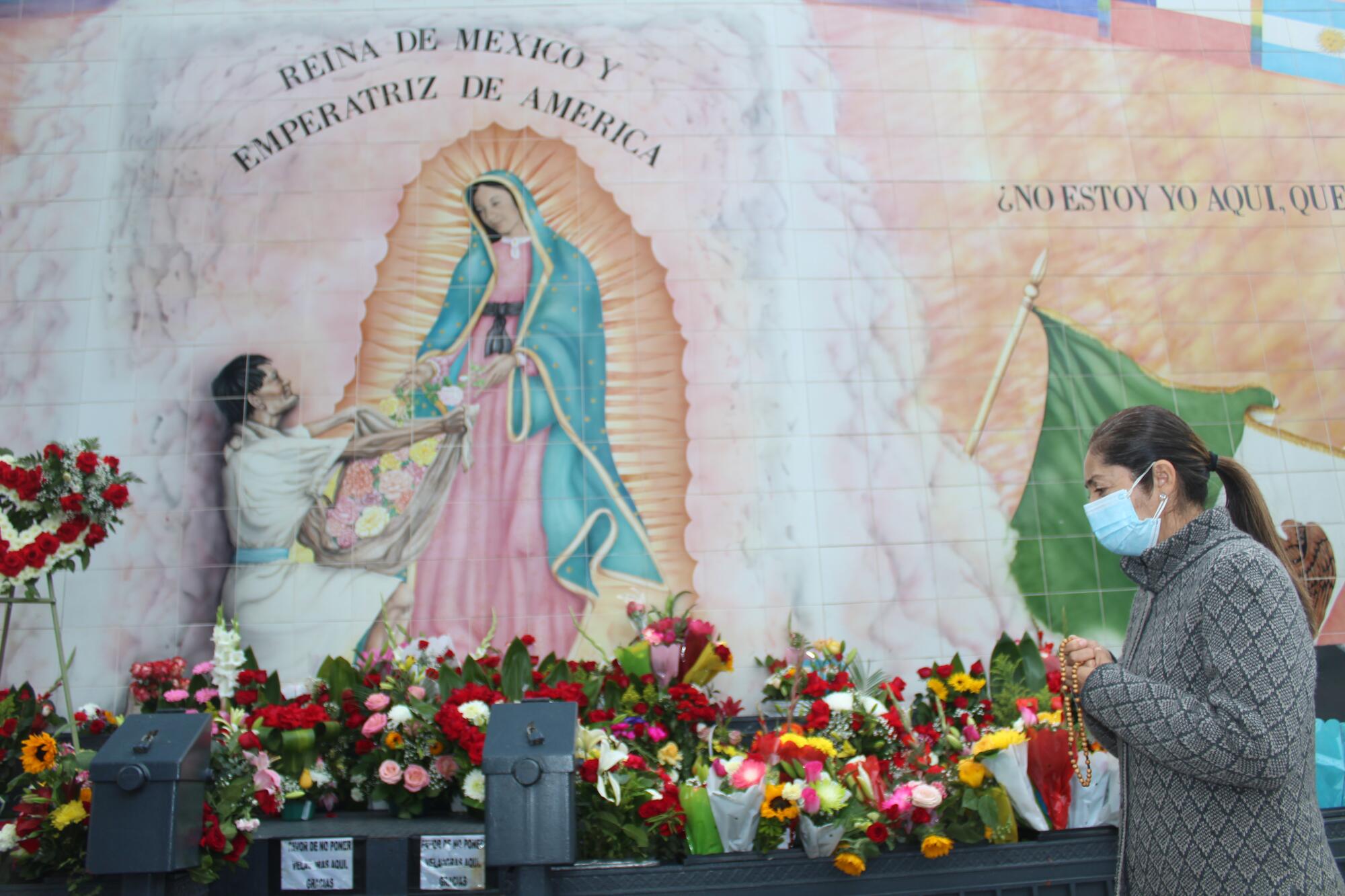 A person stands before flowers and a mural of the Virgen de Guadalupe.
