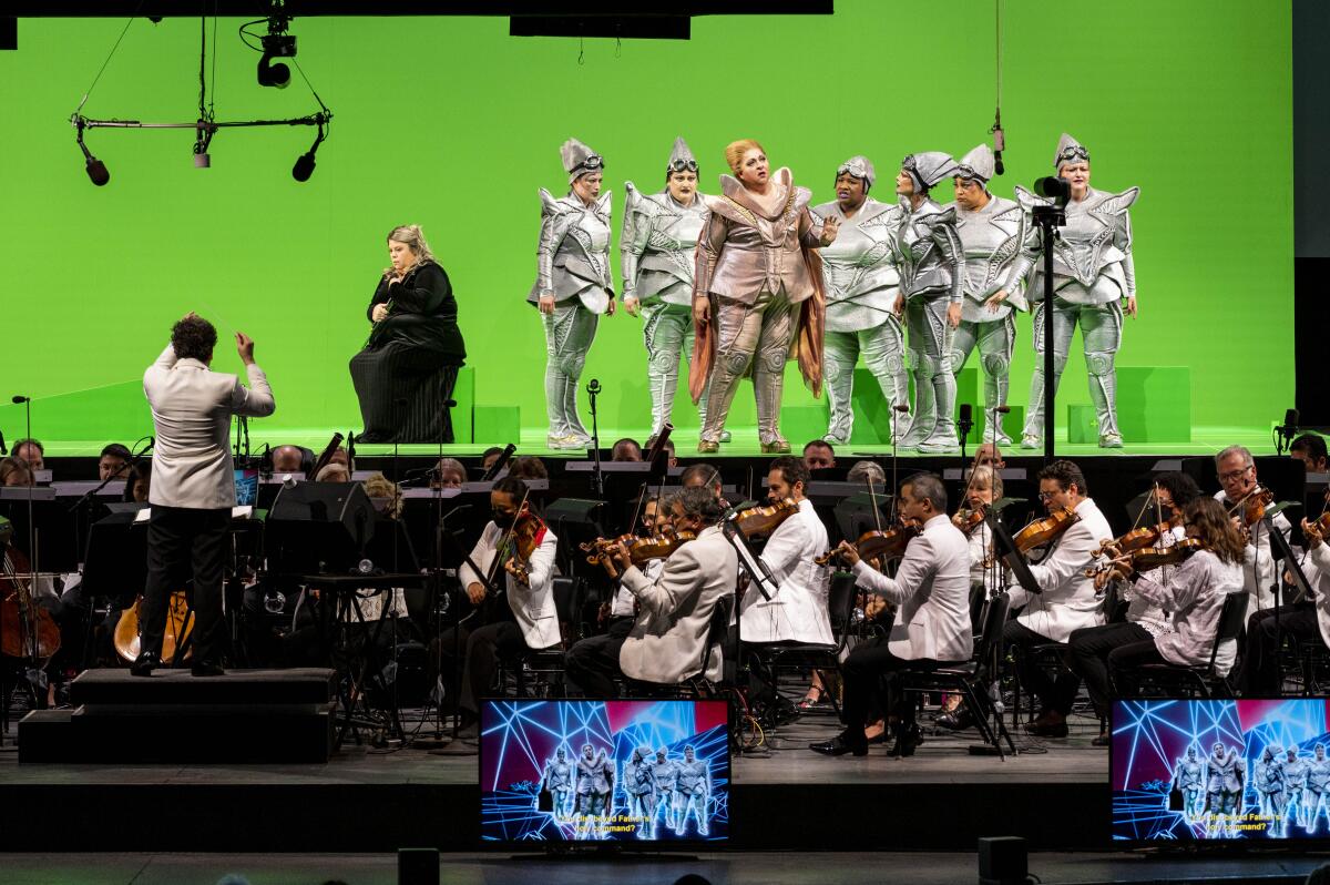 A shallow stage with a green screen, beyond an onstage orchestra.