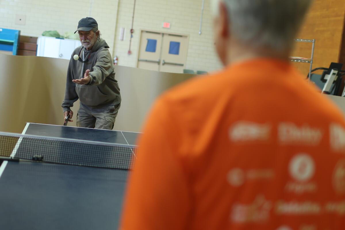 A man in a sweatshirt and ballcap, seen over the shoulder of another person, prepares to serve a pingpong ball