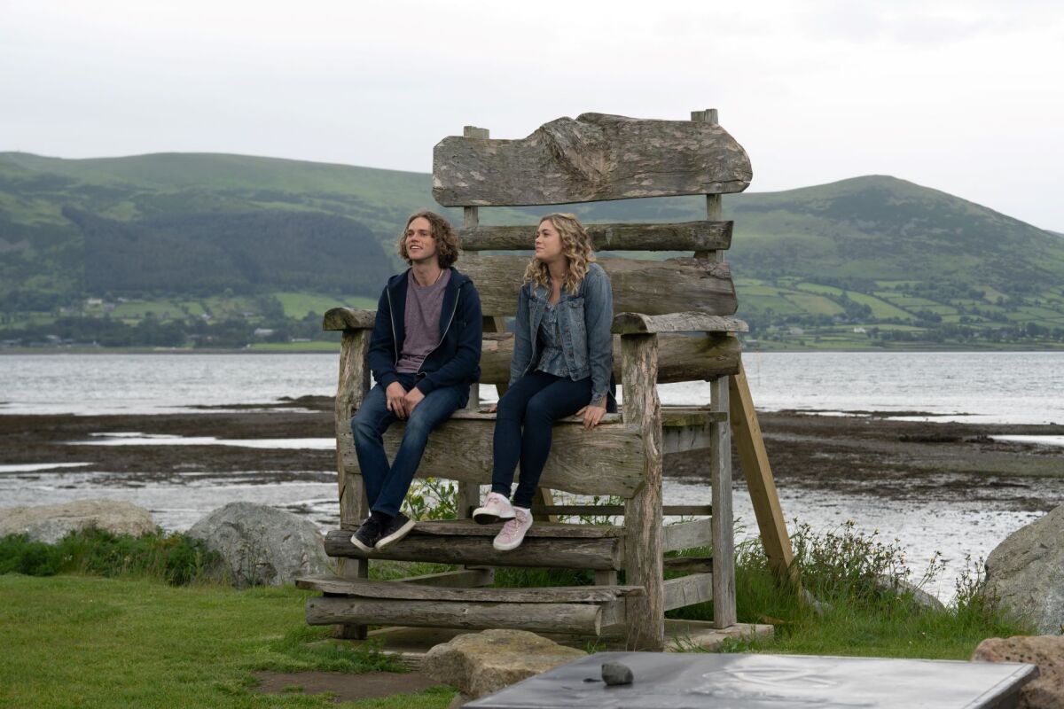 Jedidiah Goodacre and Rose Reid sit on a giant wooden chair in a scene from the film "Finding You."