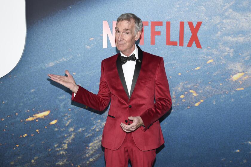 A man in a bright red velvet tuxedo gestures with one arm upon arrival at an event