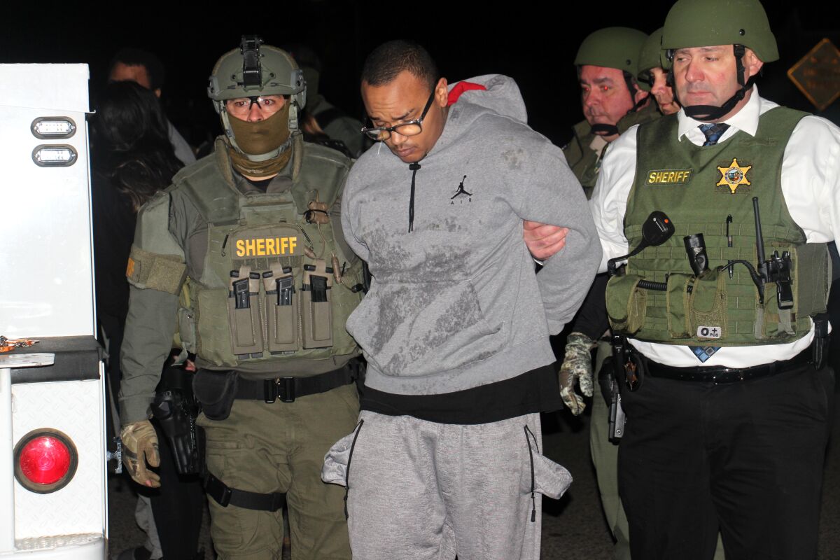 Sheriff's deputies lead a handcuffed man wearing a gray track suit and glasses. 