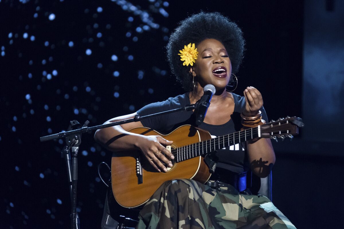 A seated woman holding an acoustic guitar and singing into a microphone