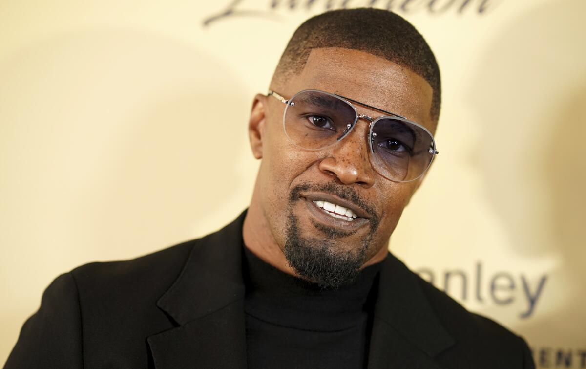 Jamie Foxx smiles and tilts his head while wearing aviator sunglasses and a black shirt and suit