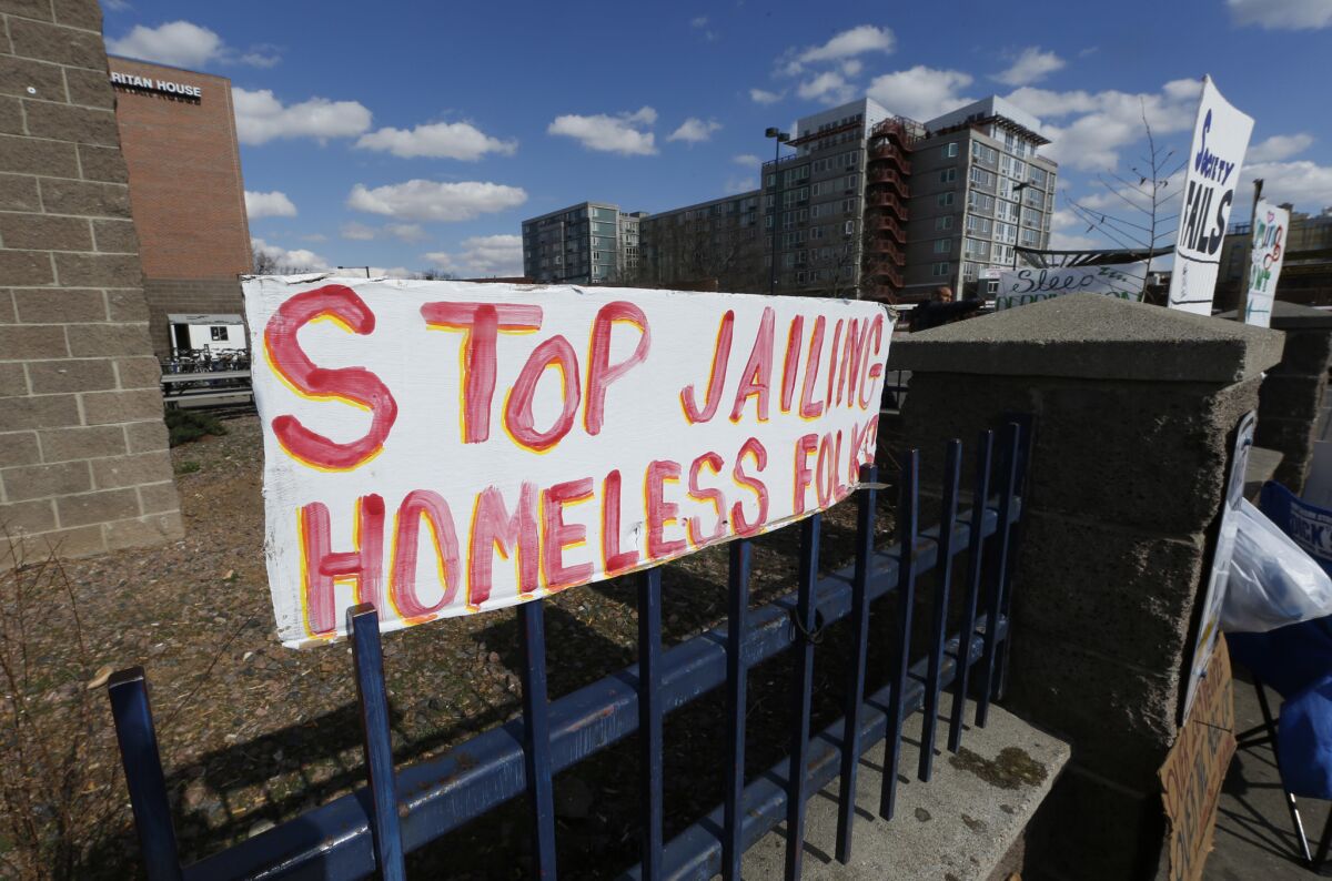 A sign hangs on a fence around the Samaritan House in Denver as homeless people from a camp are moved by the city. A new high-rise apartment building can be seen in the background.