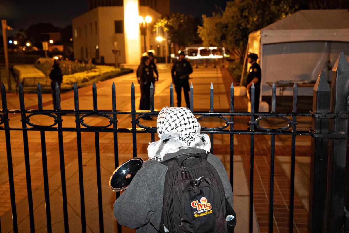 A protester with a backpack and kaffiyeh standing at a gate to USC's campus, with police officers in background