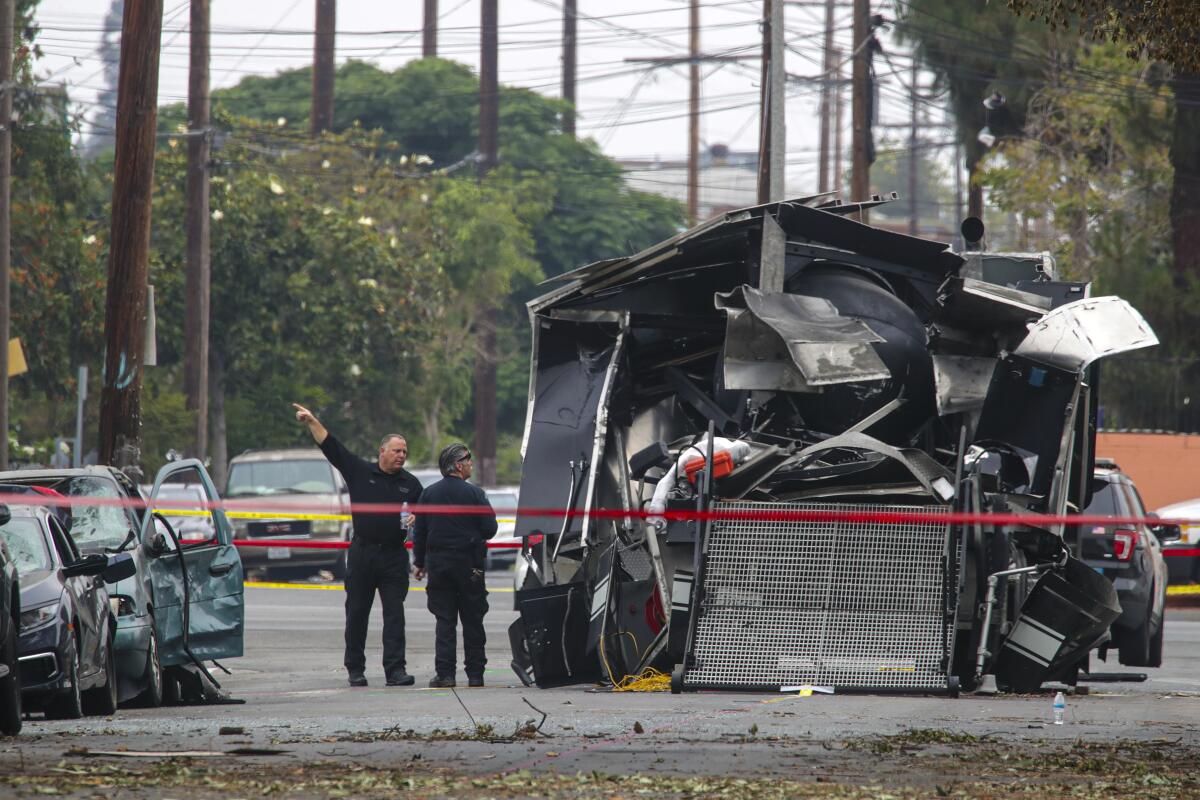 Two men, one pointing, stand alongside the twisted wreckage of a vehicle. 